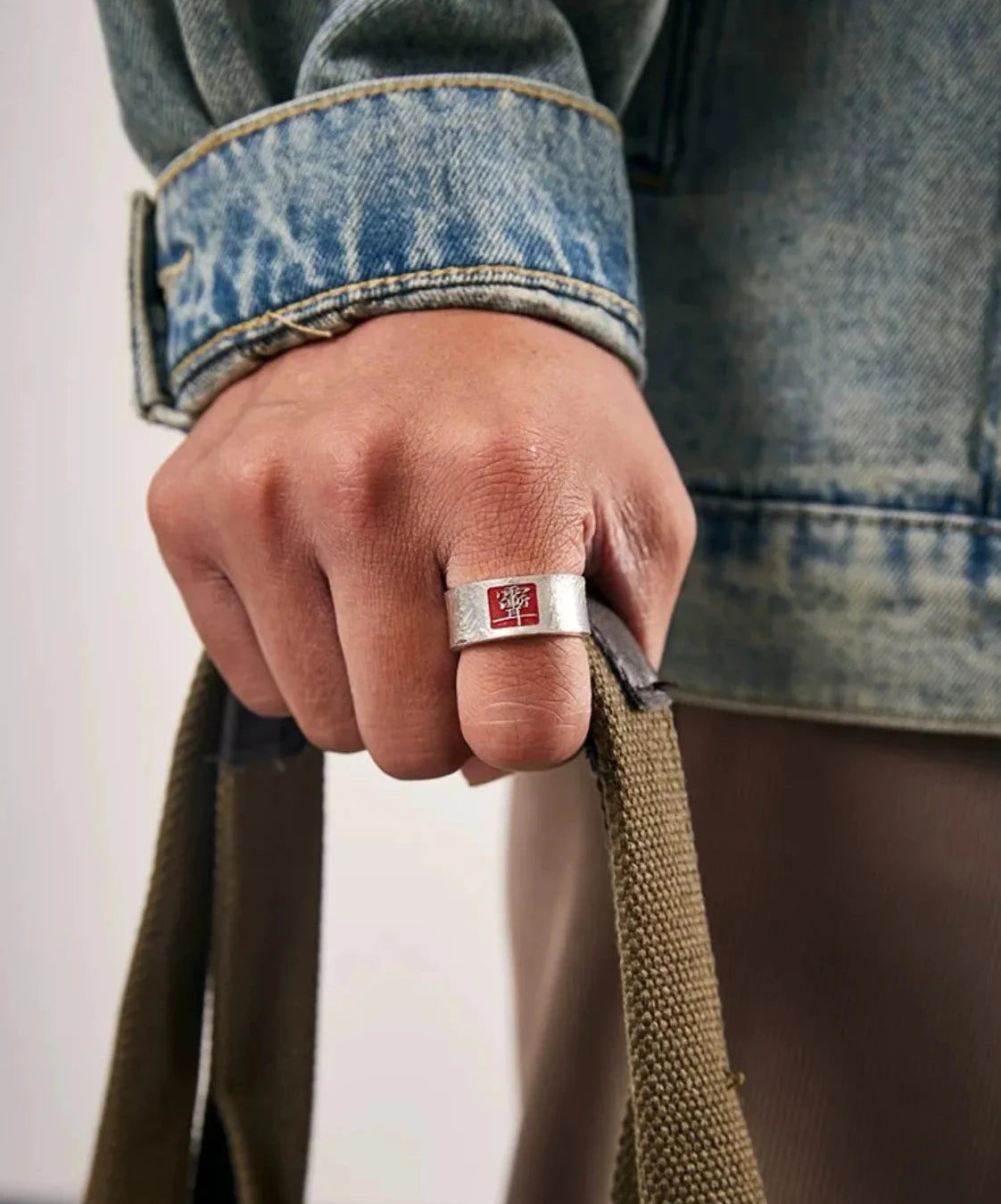 A casual shot of a hand holding a bag, showing how the silver and red ring complements a blue denim jacket for a modern streetwear look
