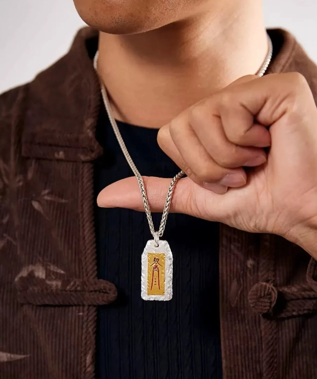 Close-up of a hand holding the oriental wealth charm pendant, highlighting the vermilion calligraphy against a brown jacket