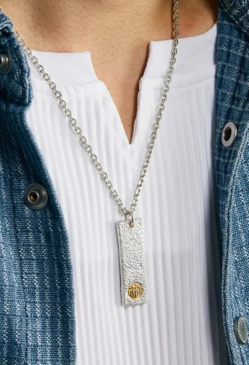 Close-up of a handcrafted oxidized silver bar pendant featuring a twisted gold accent, worn on a simple cord with earth-tone vintage clothing