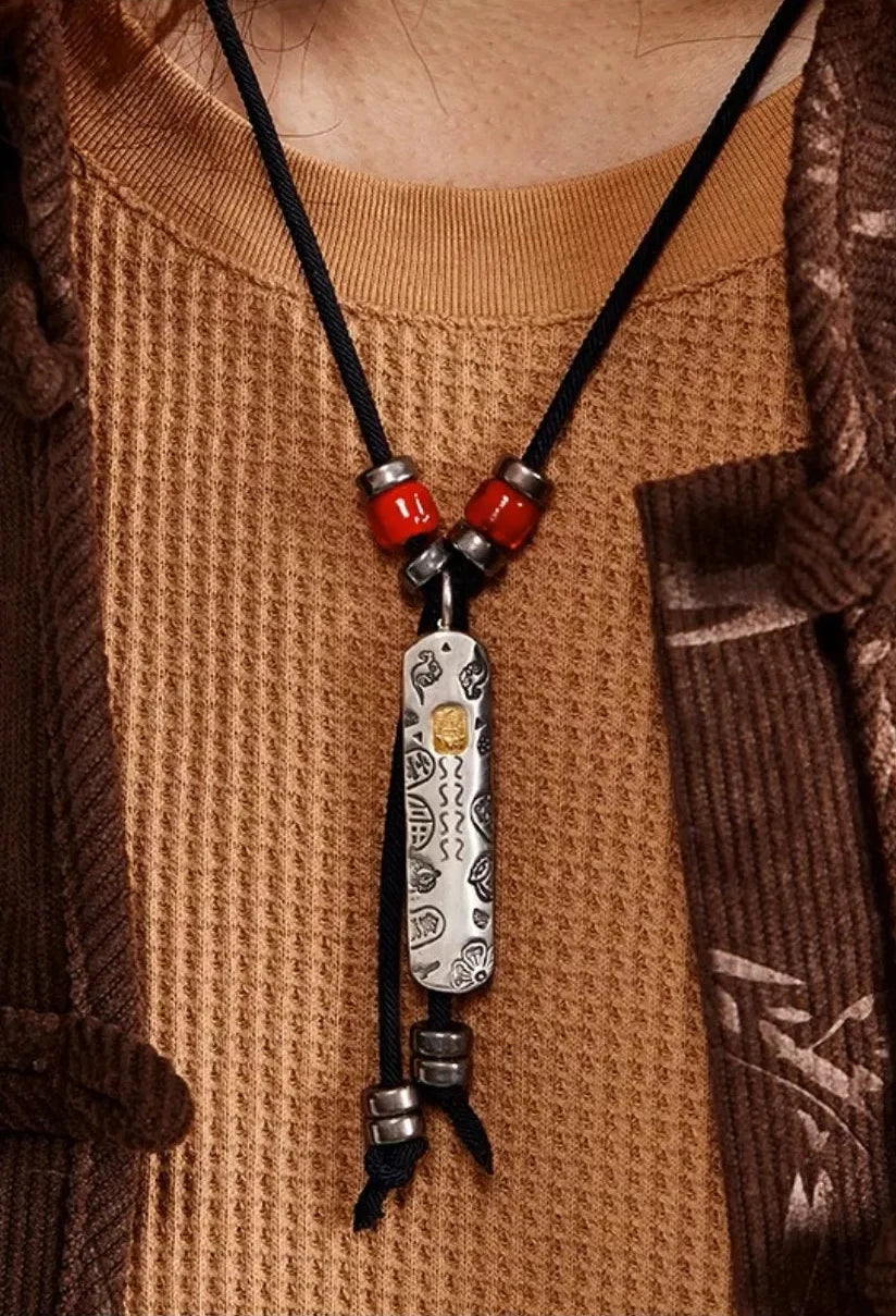 Close-up of a tribal style engraved silver bar pendant featuring red ceramic beads on a black cord, worn with earth-tone layered clothing
