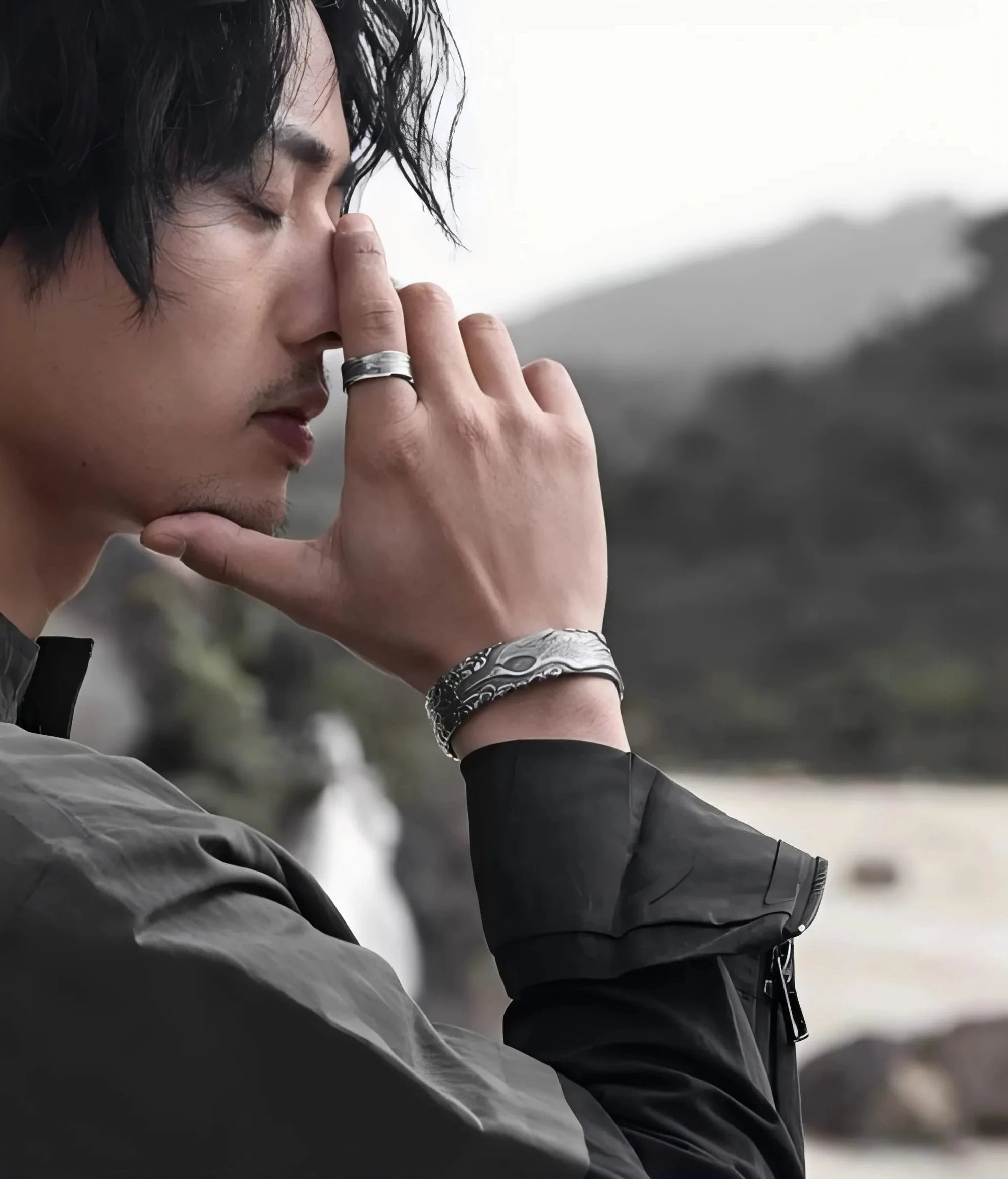 A moody portrait of a model in dark techwear, wearing the crane bangle while in a contemplative pose against a rocky background