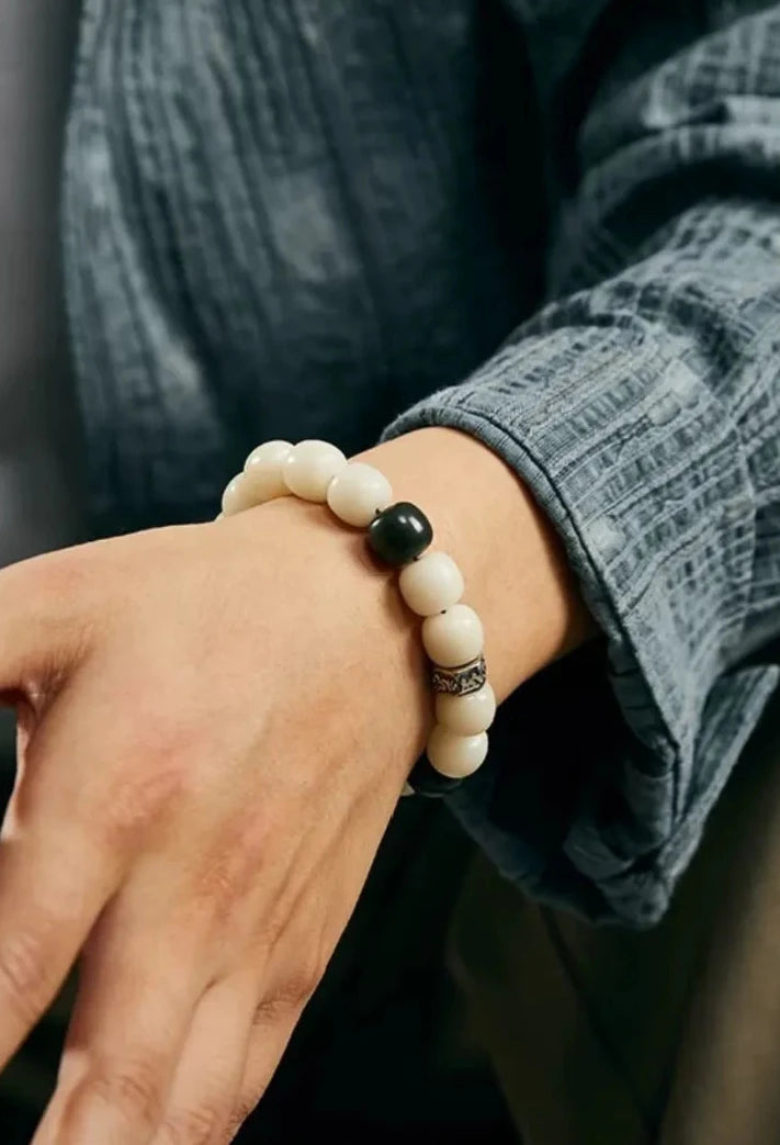 Close-up on the wrist showing the contrast between the white Bodhi, black beads, and silver charm against a textured blue linen shirt
